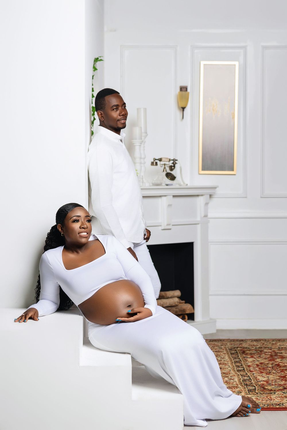 Expectant mother in a flowing white gown beside a fireplace in soft daylight