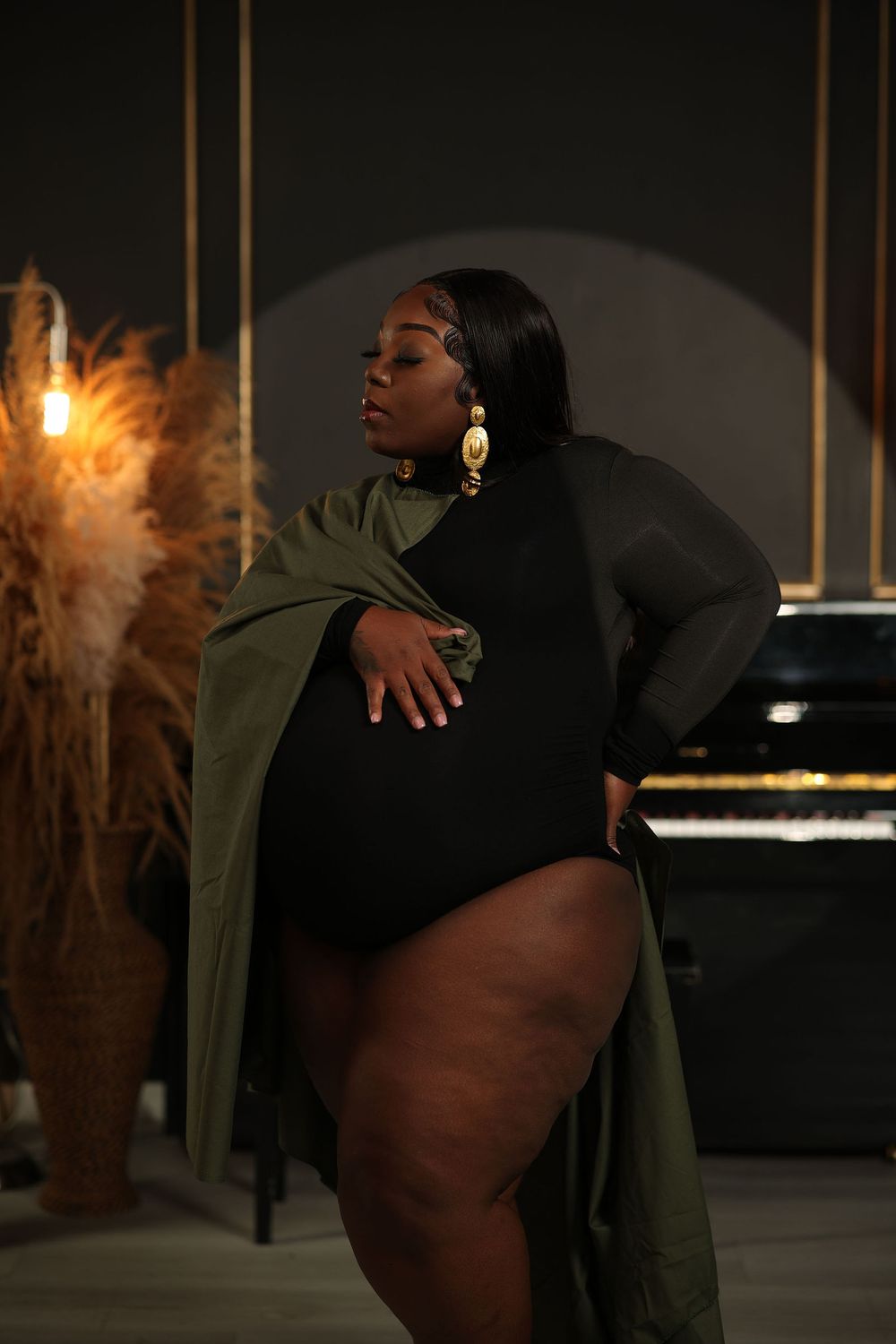 Mother to be in an olive cape beside a black piano and pampas grass