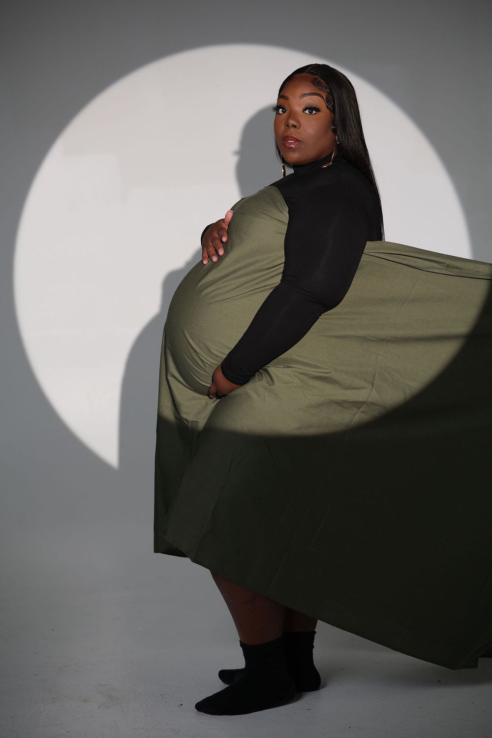 Expectant mother sweeping an olive cape across a disc of studio light