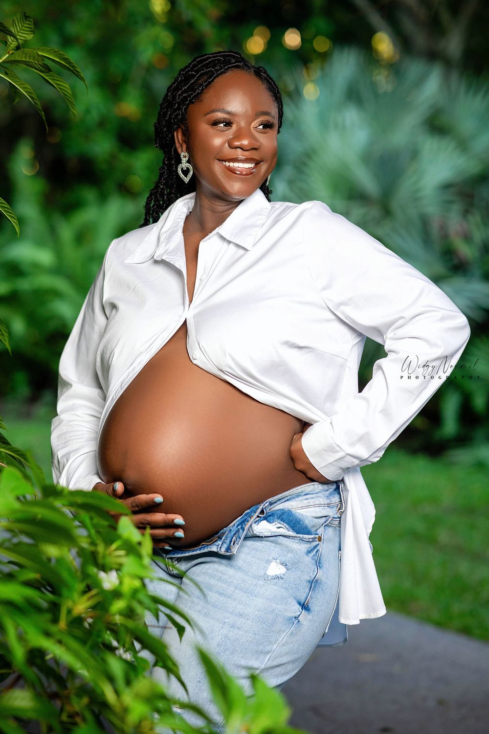 Expectant mother in an open white shirt holding her bare belly in a tropical garden