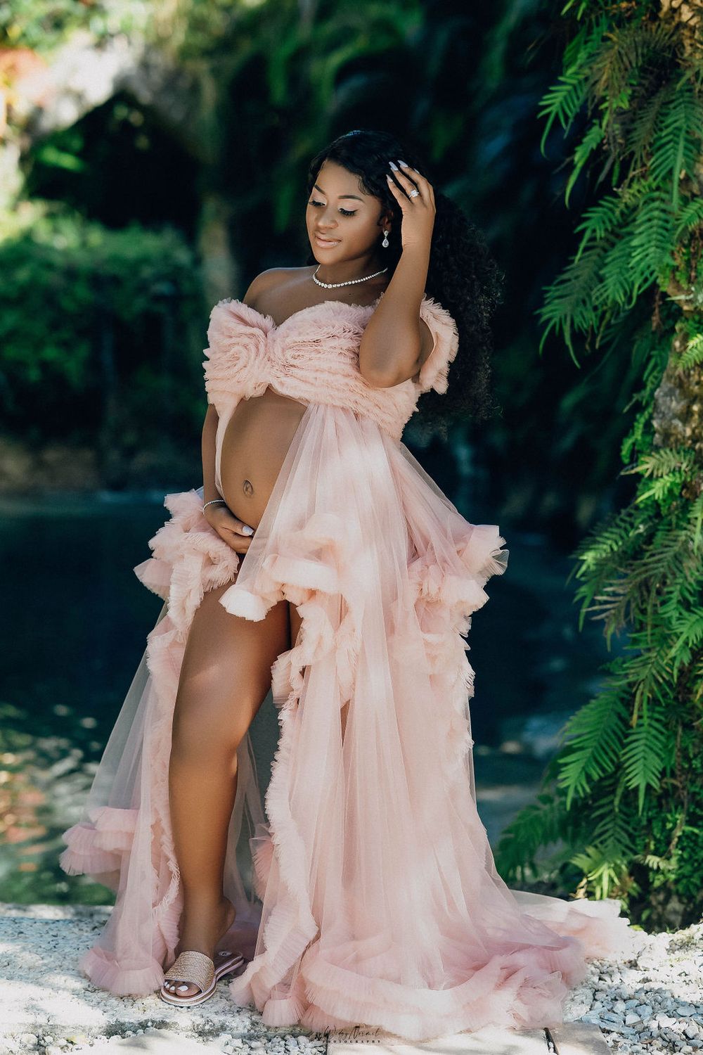 Expectant mother in a blush gown among the ferns at the water's edge