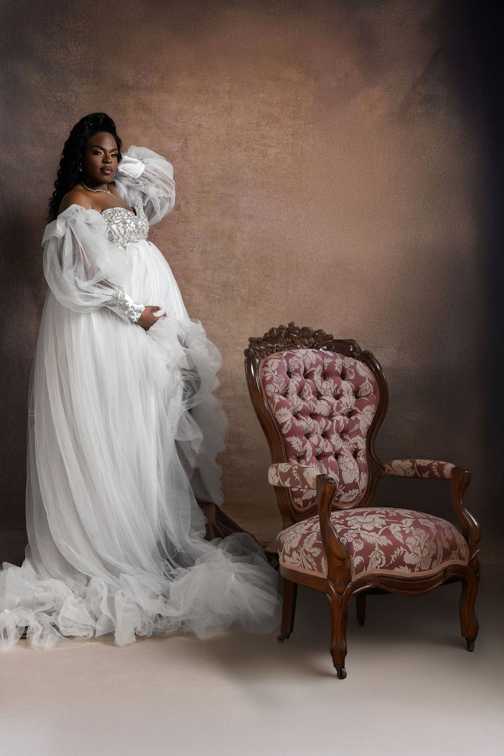 Mother to be in a white tulle gown beside an antique chair in the studio