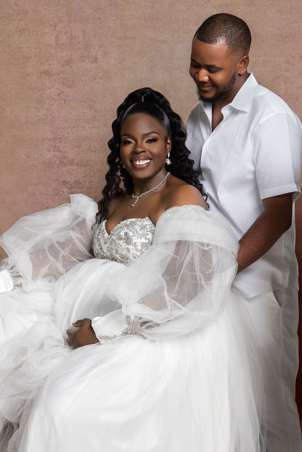 Expecting couple seated together, her white tulle gown spilling across the floor