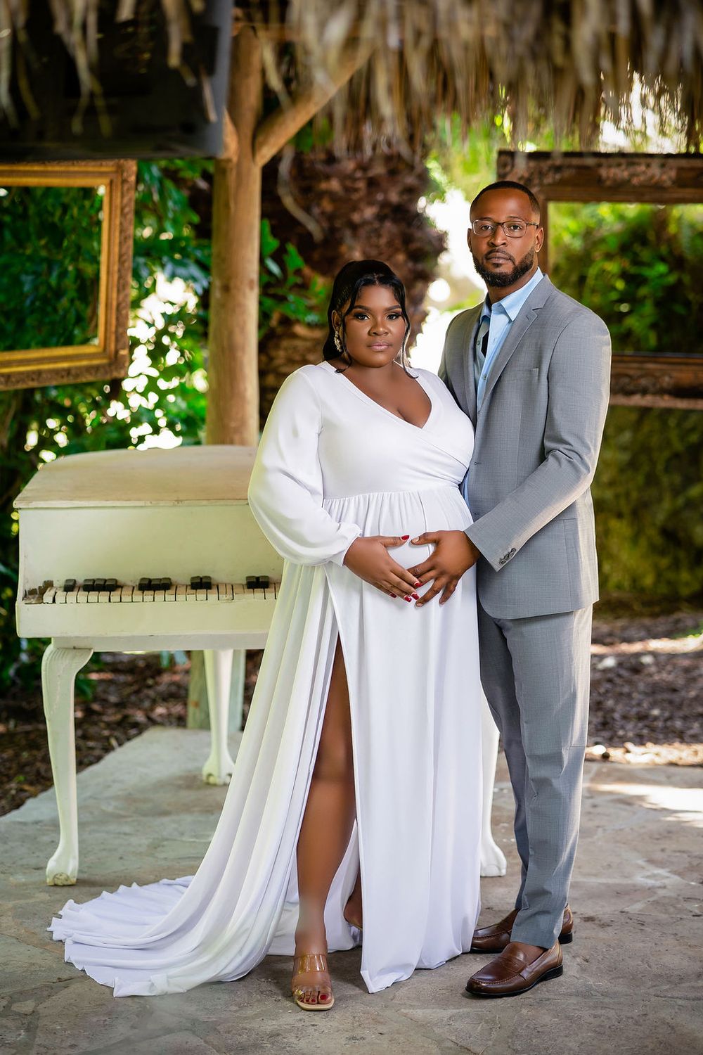 Expecting couple beside a white piano in a tropical garden, his hand on her belly