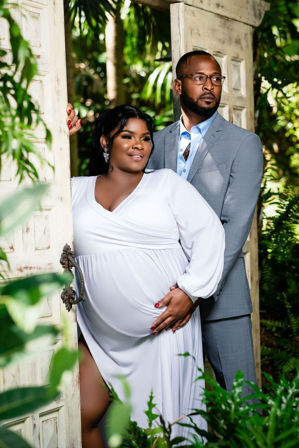 Expecting couple in a white gown and grey suit among the ferns