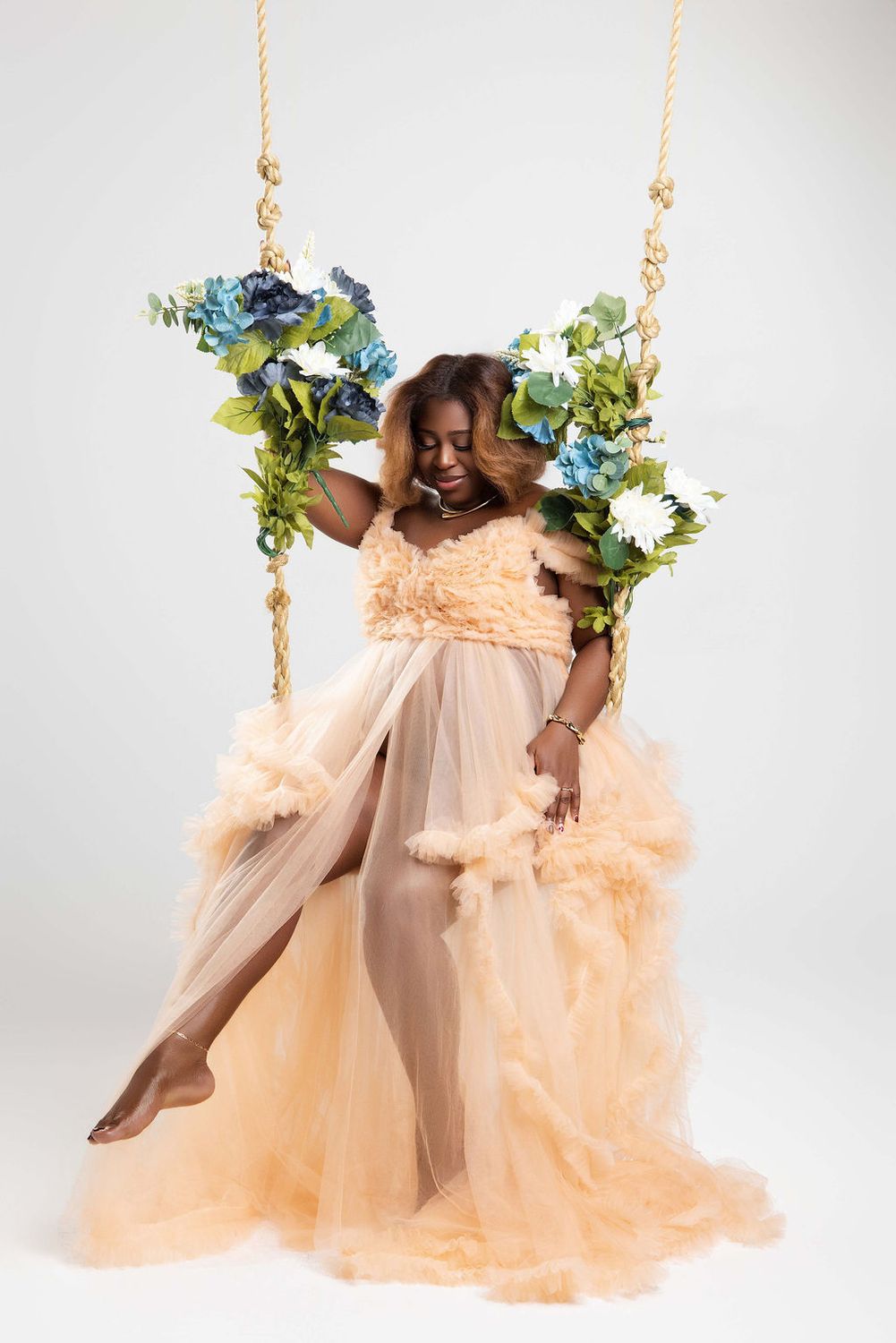 Expecting mother on a flower swing wearing peach tulle, hands on her belly