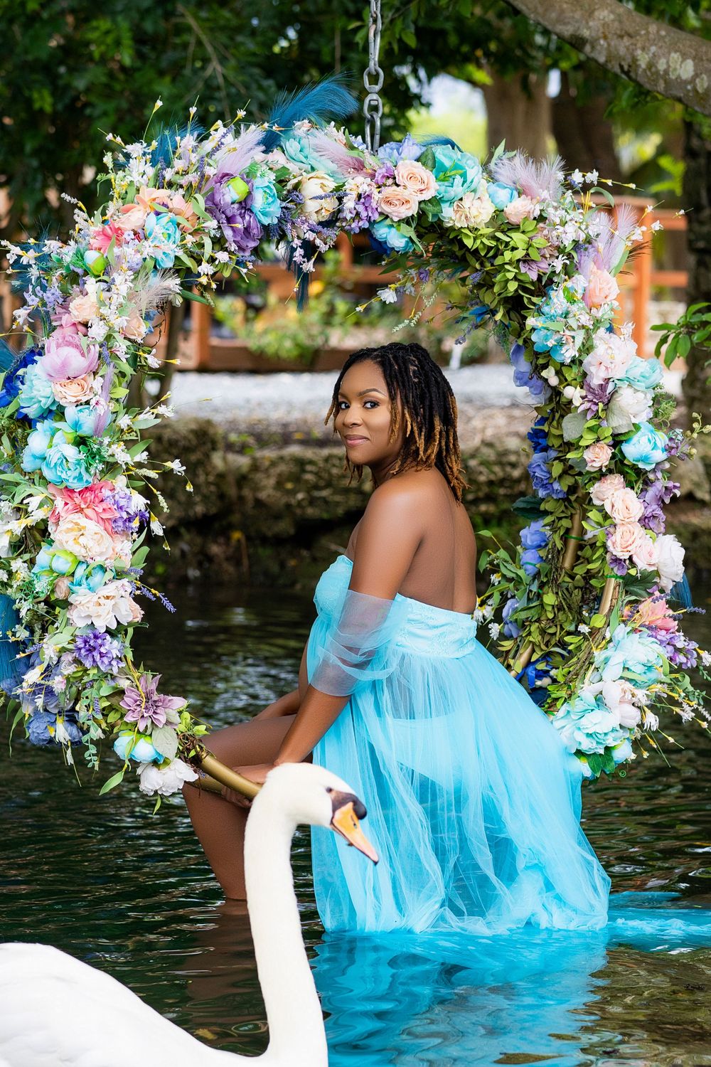 Mother to be in a blue gown on a floral swan arch above still water