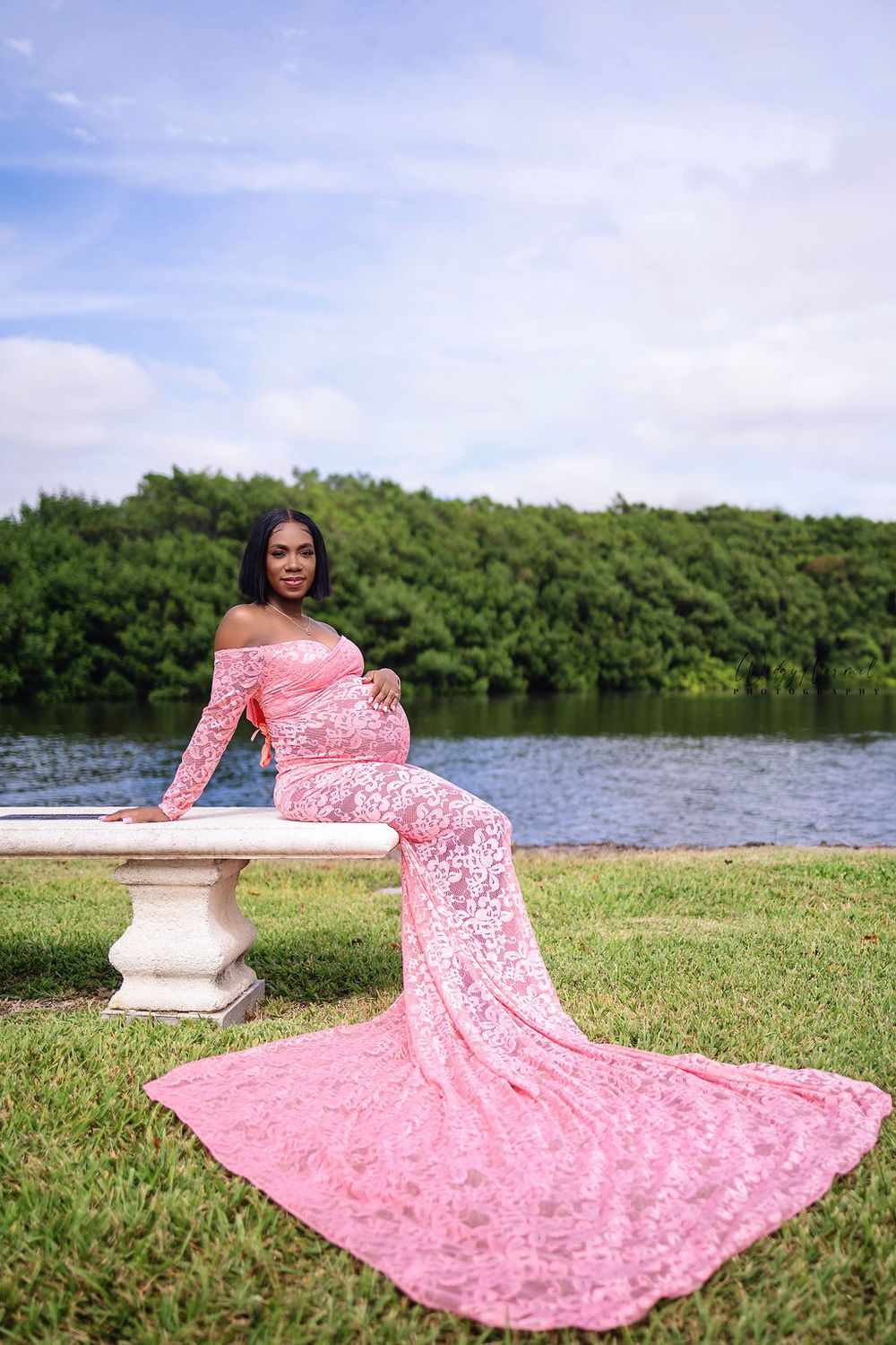 Mother to be in a pink lace gown seated on a stone bench by the water