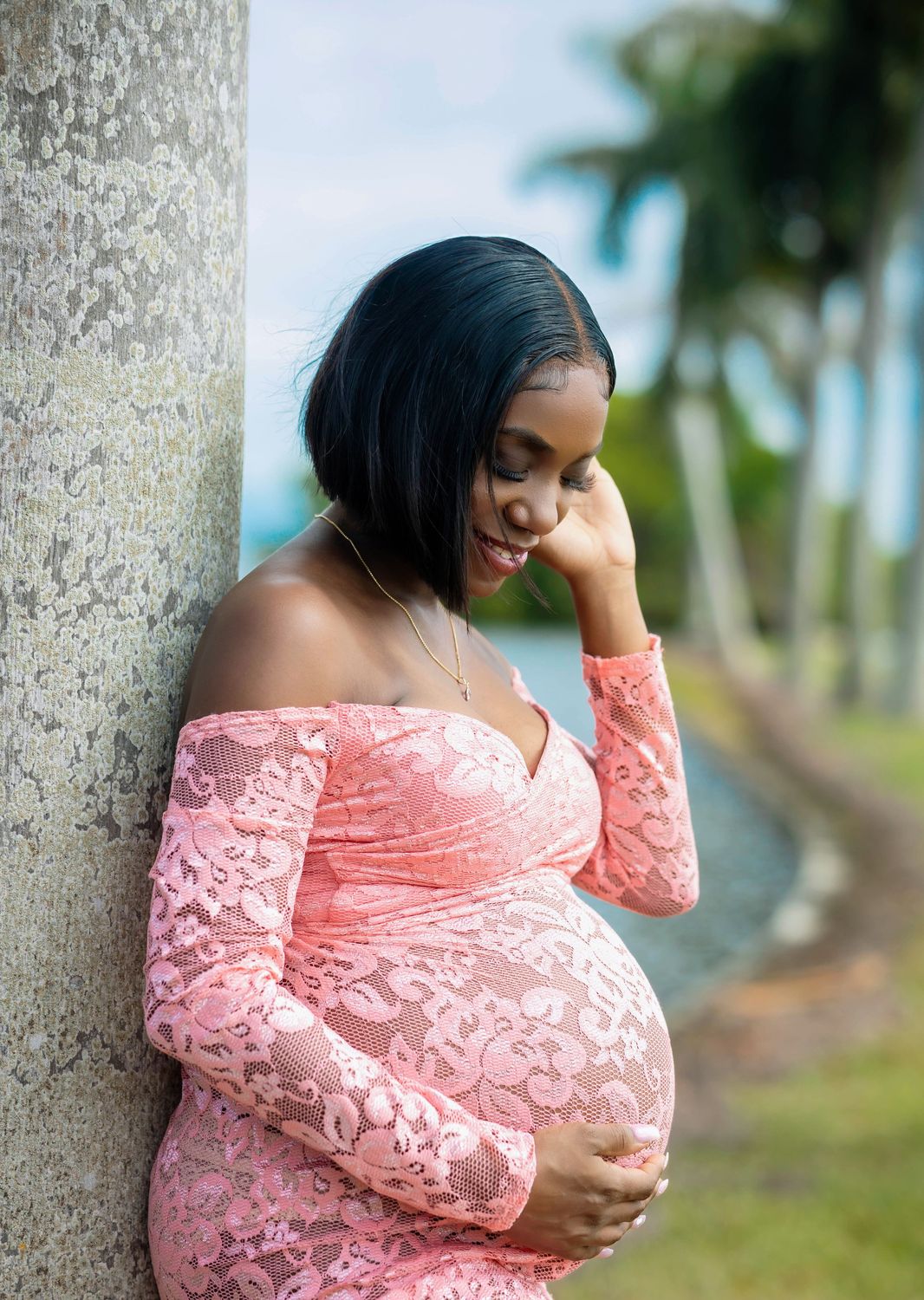Expectant mother in a blush lace gown resting her hands on her belly in soft South Florida daylight