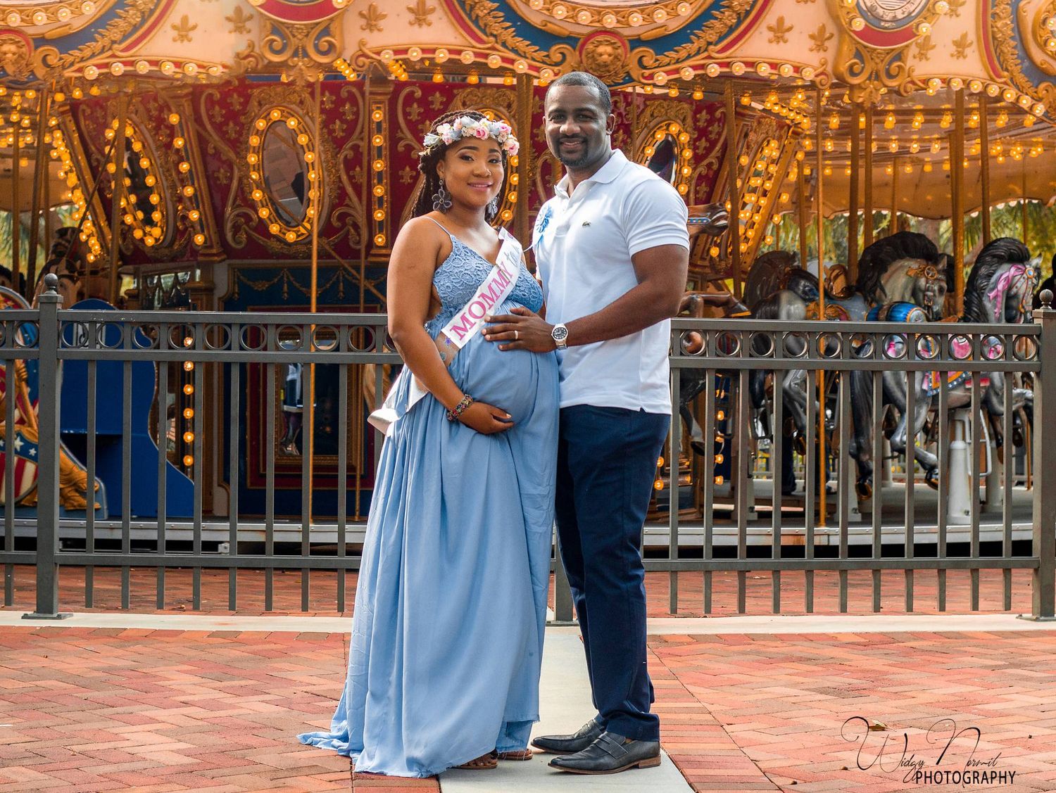 Expecting couple in front of a lit carousel, the mother to be in a blue gown and a mommy to be sash