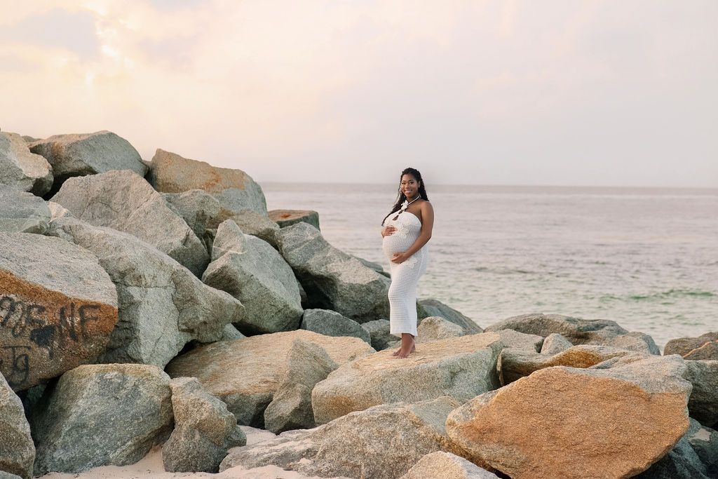 Expectant mother in white standing among the breakwater rocks at a South Florida beach