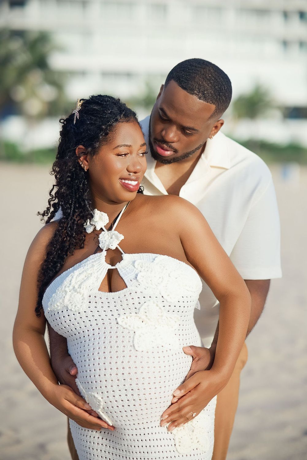 Mother to be in a white crochet dress cradling her belly at the beach