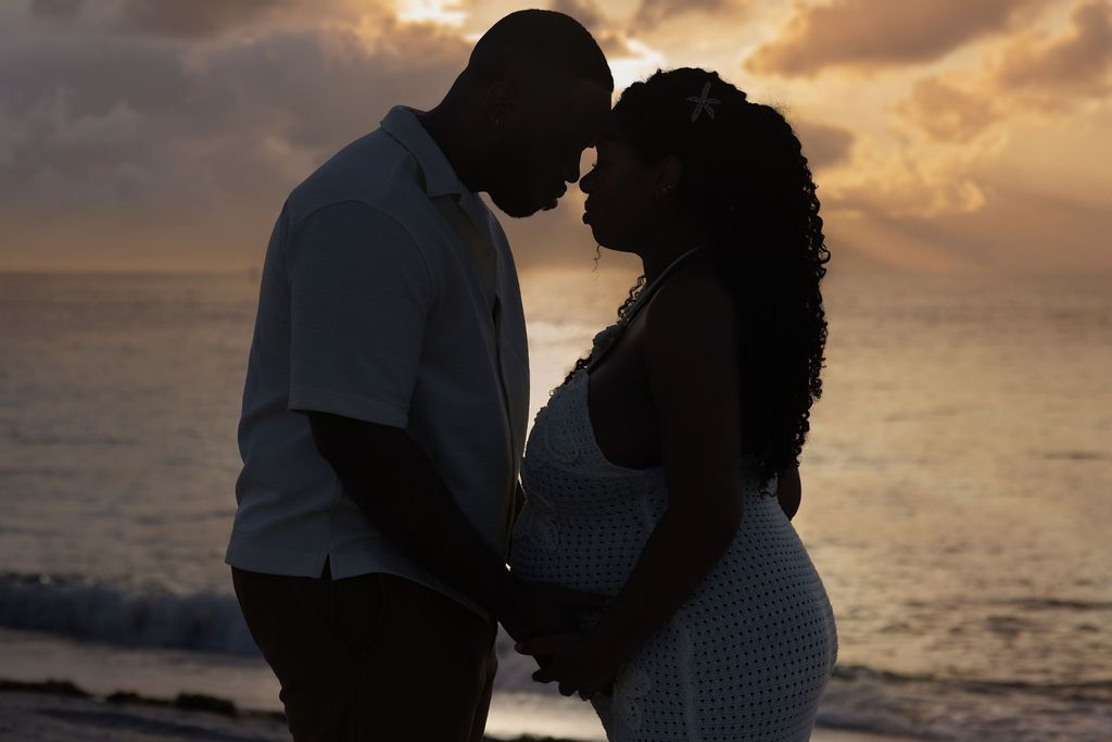 Expecting couple silhouetted against a gold and lilac sky at the water's edge