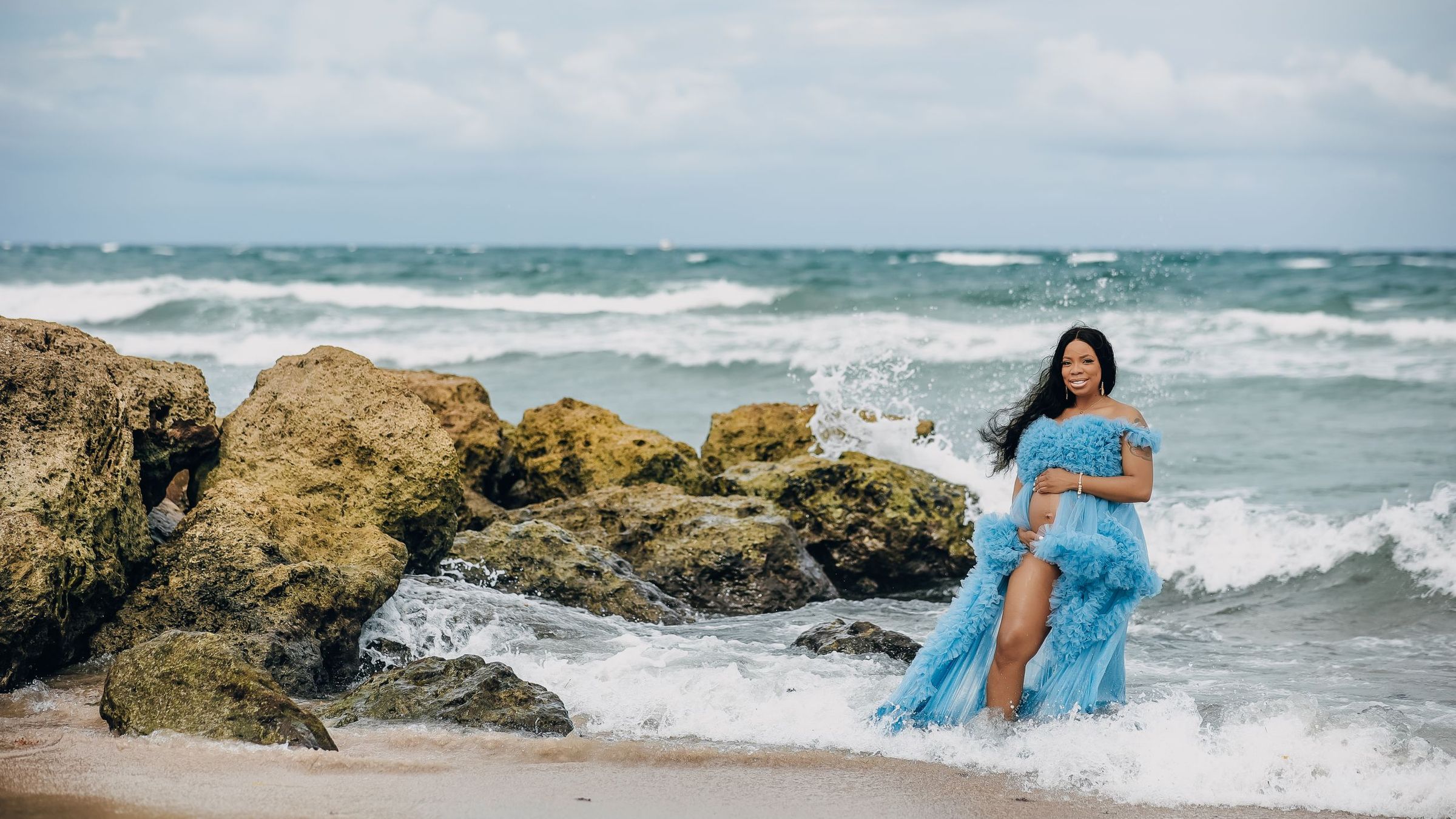Expectant mother in a powder blue tulle gown on the ocean rocks as the surf breaks