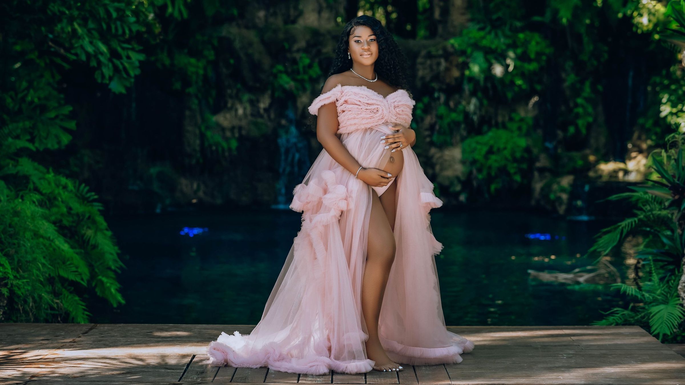 Expectant mother in a blush ruffled gown beside a hidden blue lagoon in Miami